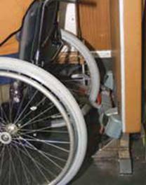 Close up photograph of manual wheelchair user in wheelchair securement area on high floor Muni bus with wheelchair clamp and seatbelt applied.