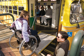 Photograph of woman in manual wheelchair using wayside platform and bridge plate to board Muni historic streetcar.  Streetcar operator is shown assisting her in boarding. 