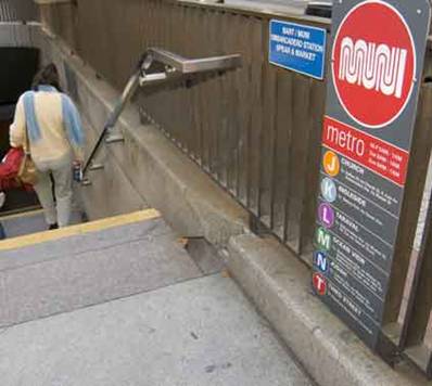 Photograph of staircase to shared BART / Muni Metro subway station that shows the visual and tactile signage located adjacent to the top of the stairs and handrail.