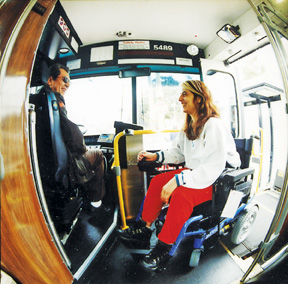 Photograph of woman in power wheelchair boarding Muni high floor bus via a wheelchair lift. She and the bus operator are smiling at each other.